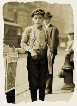 Newsboy holding newspapers and standing next to a paper stand on a commercial street, 1904